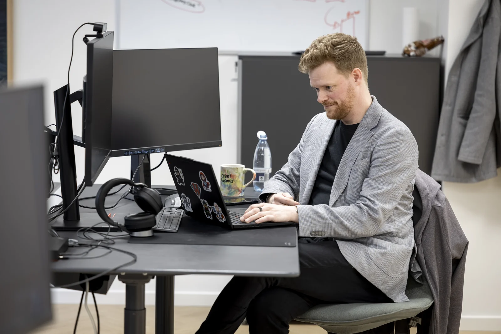Sekvens-konsulent i grå ternet blazer sidder fokuseret ved sit skrivebord og arbejder på en bærbar med stickers.